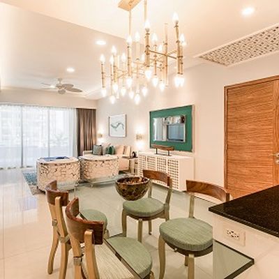 Modern dining area with glass table, wooden chairs, and a chandelier. Living room visible with sofas, large window, and wall art.