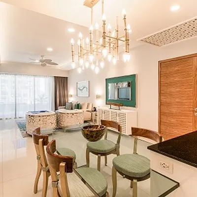 Modern dining area with glass table, wooden chairs, and a chandelier. Living room visible with sofas, large window, and wall art.