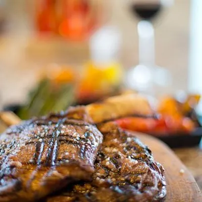 Grilled steak on a wooden board with roasted vegetables in the background, accompanied by a glass of red wine.