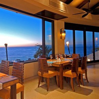 Elegant seaside restaurant interior with wooden tables and chairs, large windows showcasing a sunset view over the ocean, and warm lighting.