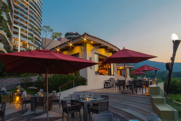 Outdoor restaurant at sunset with red umbrellas, wicker chairs, and candlelit tables. Modern building and lush greenery in the background.