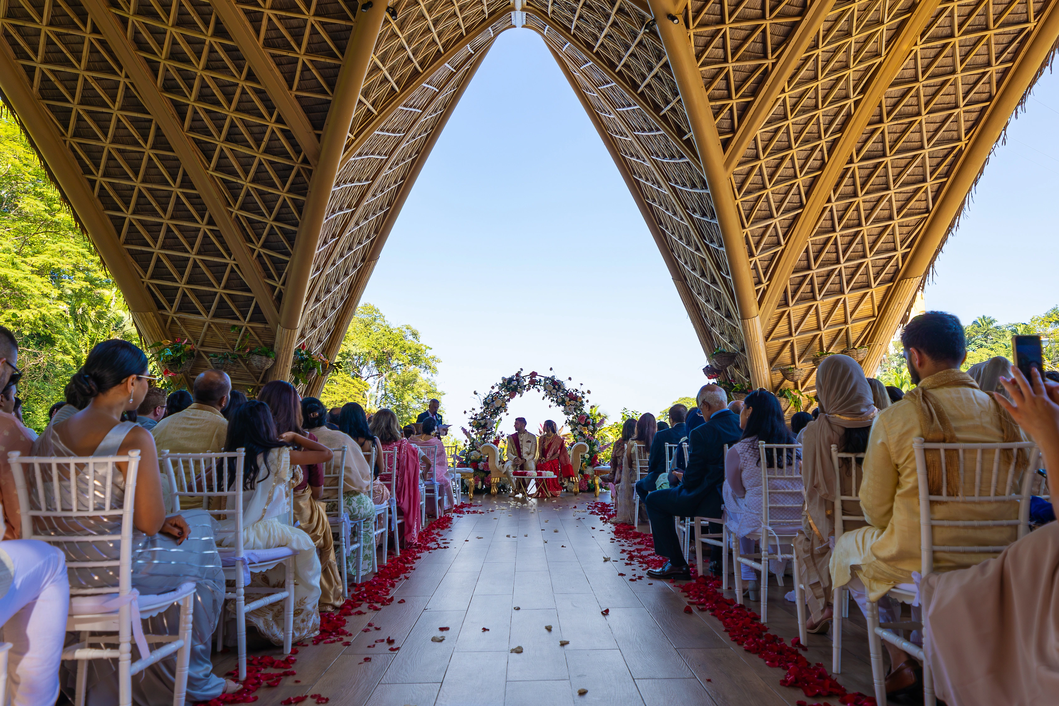 A beautiful wedding ceremony under a wooden pavilion, with guests seated and a floral arch framing the couple. The setting is outdoor and sunny.
