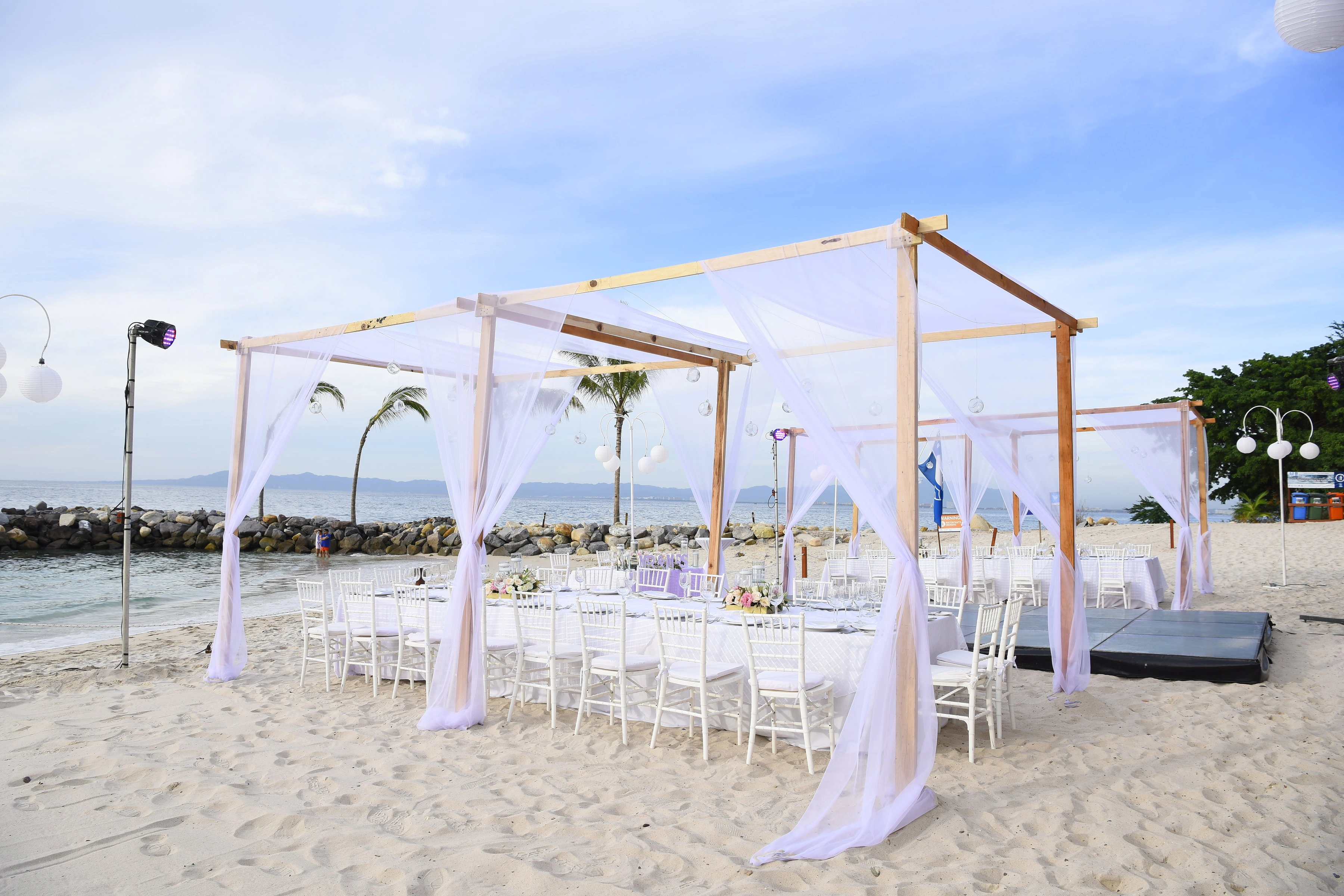 Beachfront wedding setup with white chairs and draped canopy, overlooking the ocean under a clear blue sky.