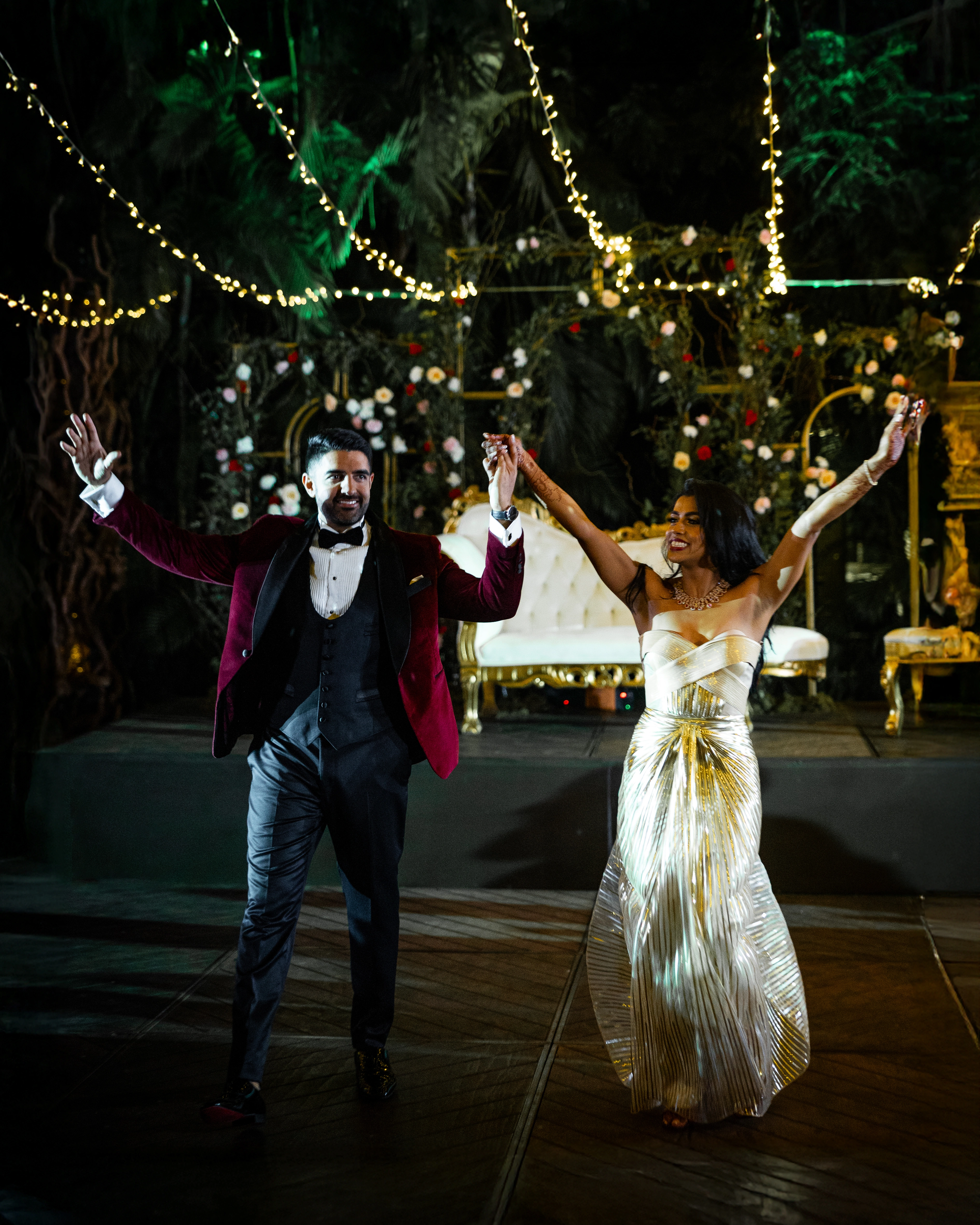 A couple in elegant evening wear smiles and raises their arms in celebration on a decorated outdoor stage with string lights.