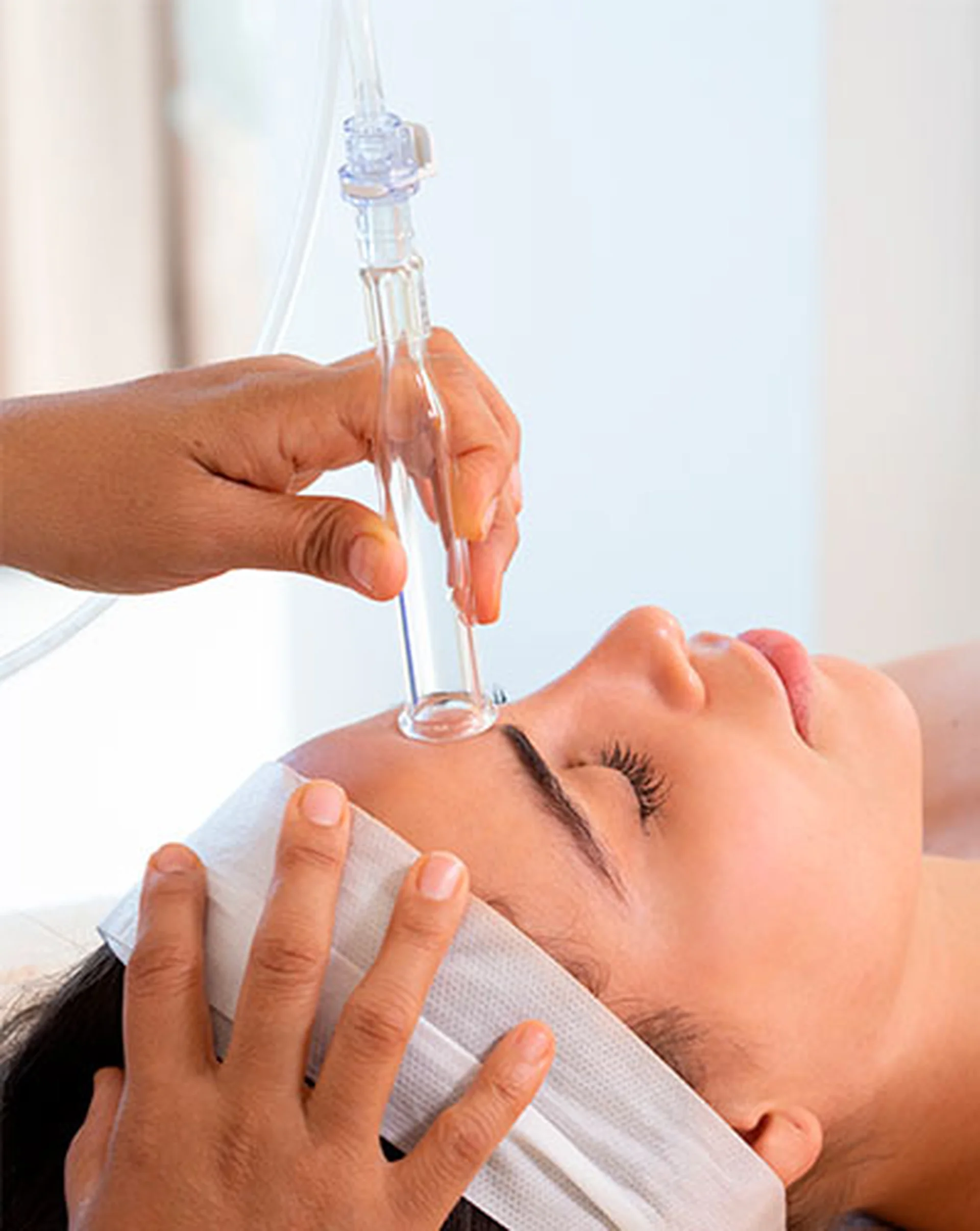 Person receiving a facial treatment with a glass suction device, lying down with eyes closed, wearing a headband.