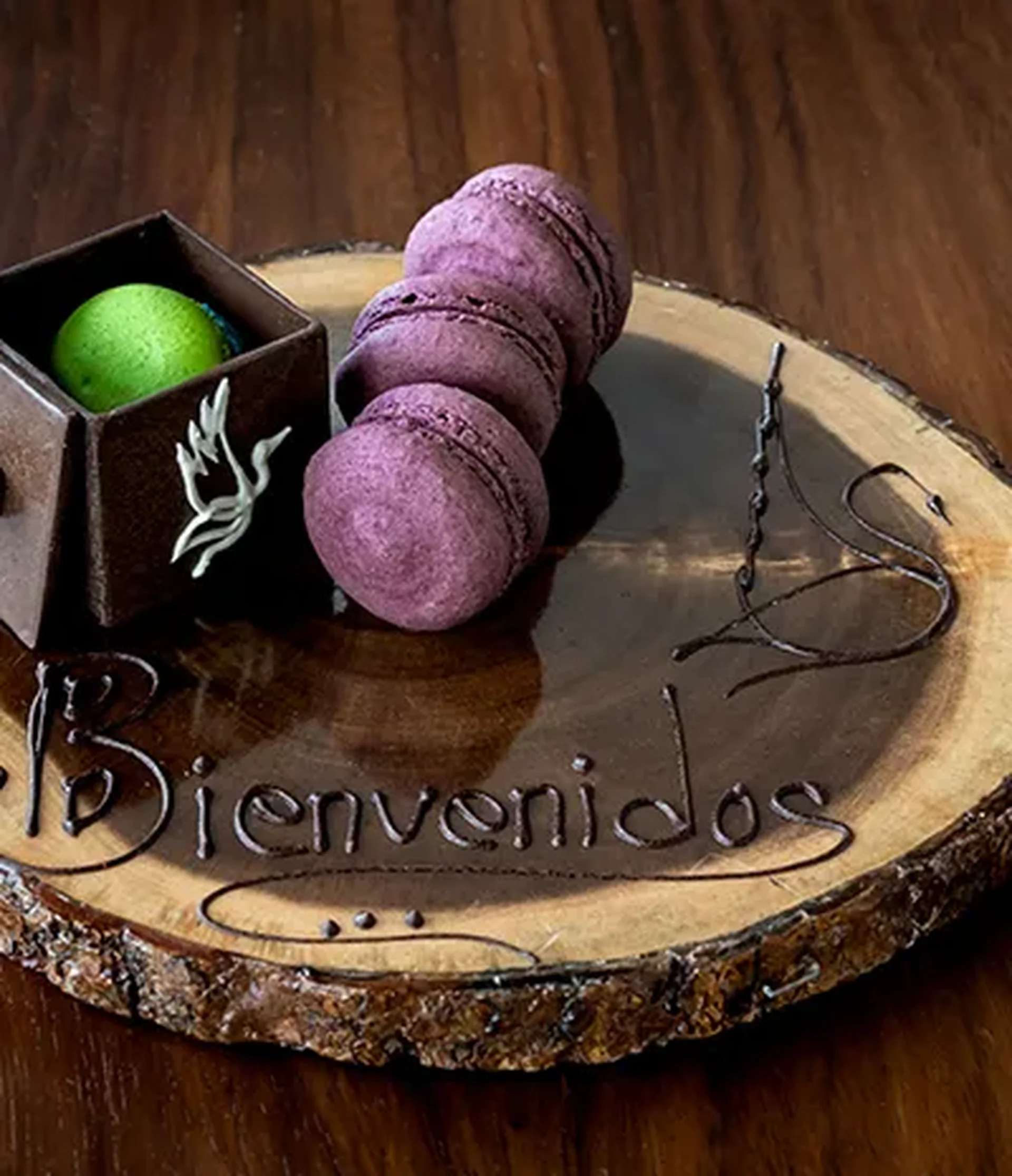 Wooden platter with purple macarons, a green sphere in a chocolate box, and "Bienvenidos" written in chocolate.