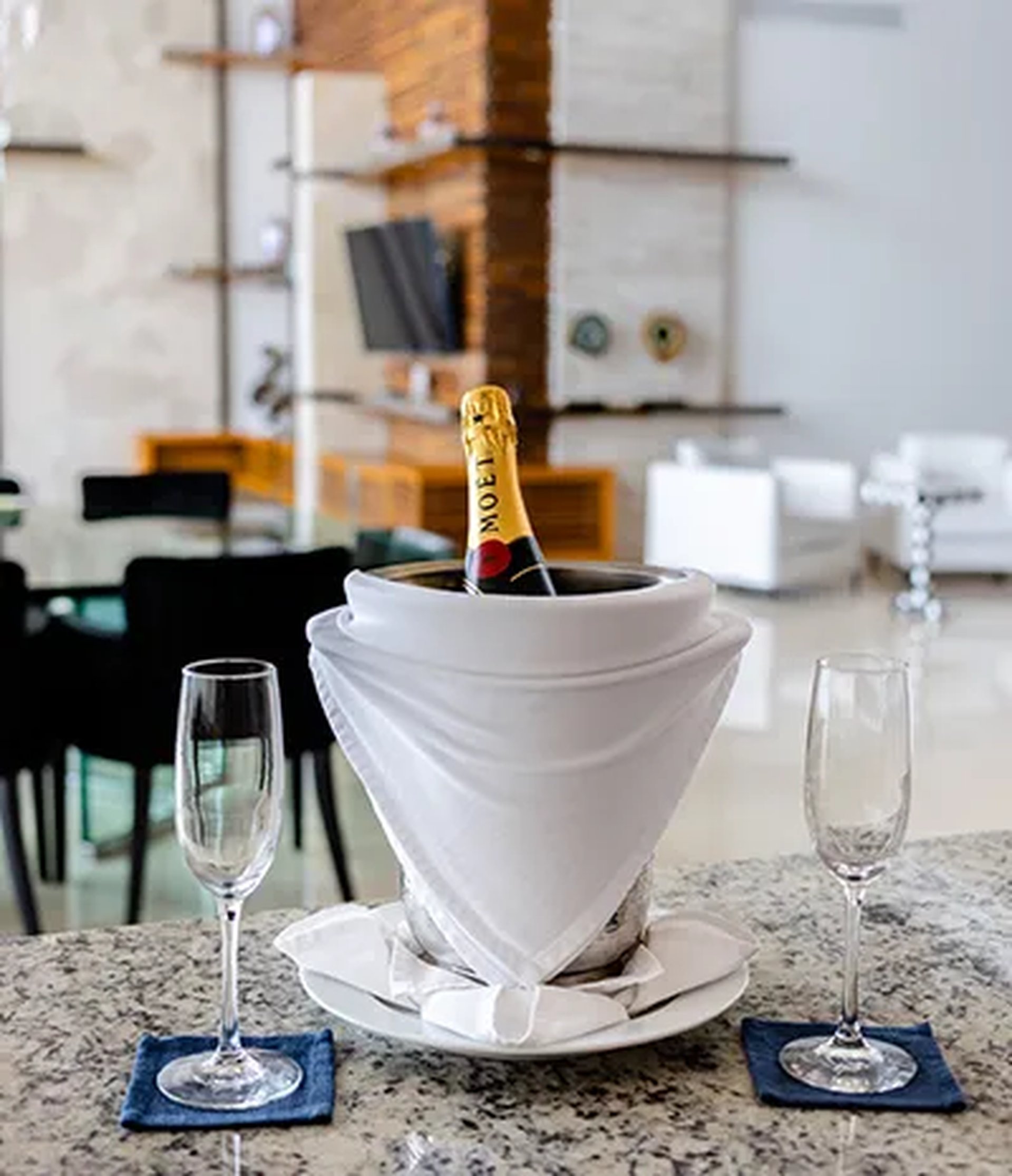 A champagne bottle in a bucket with a white cloth, flanked by two empty glasses on a granite countertop in a modern room.