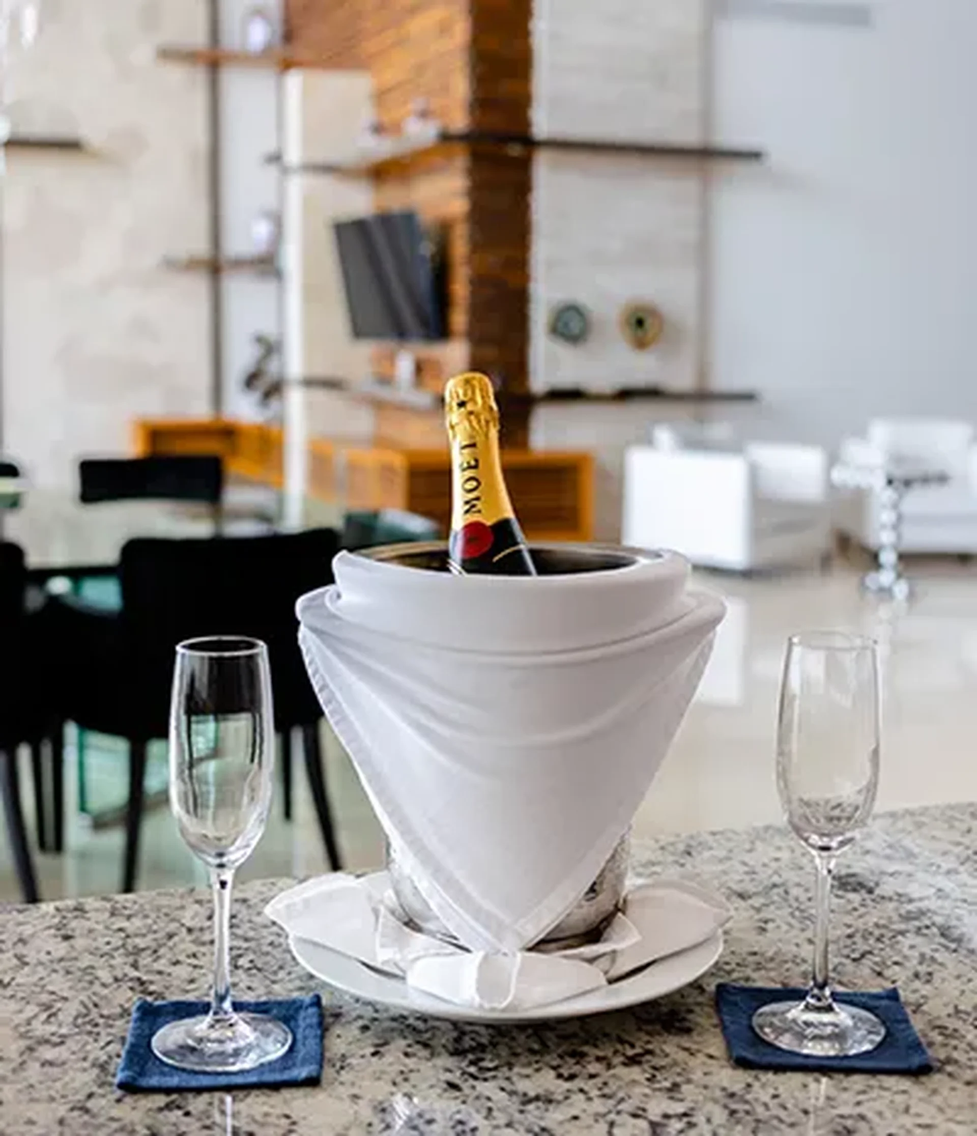 A champagne bottle in a bucket with a white cloth, flanked by two empty glasses on a granite countertop in a modern room.