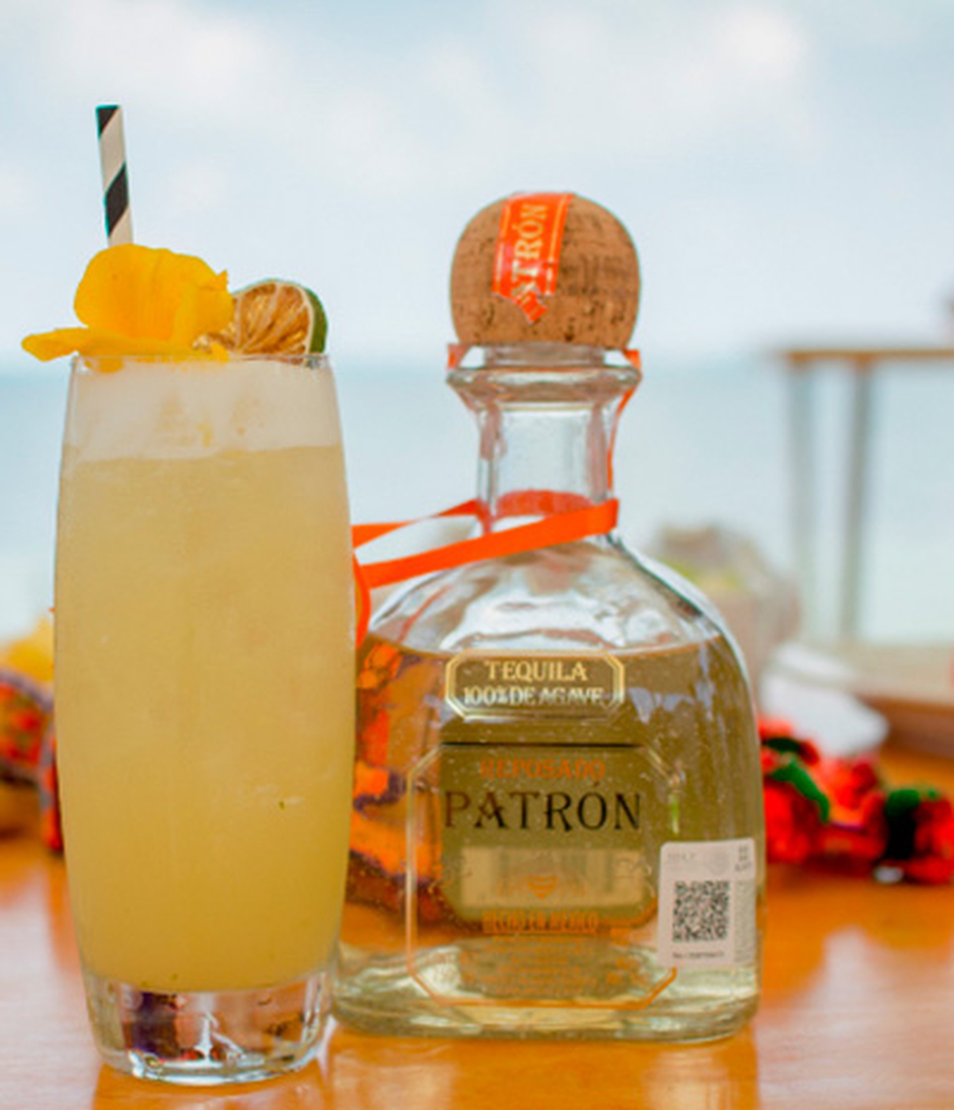 Tall cocktail with lime and pineapple garnish beside a bottle of Patron tequila on a sunny beachside table.