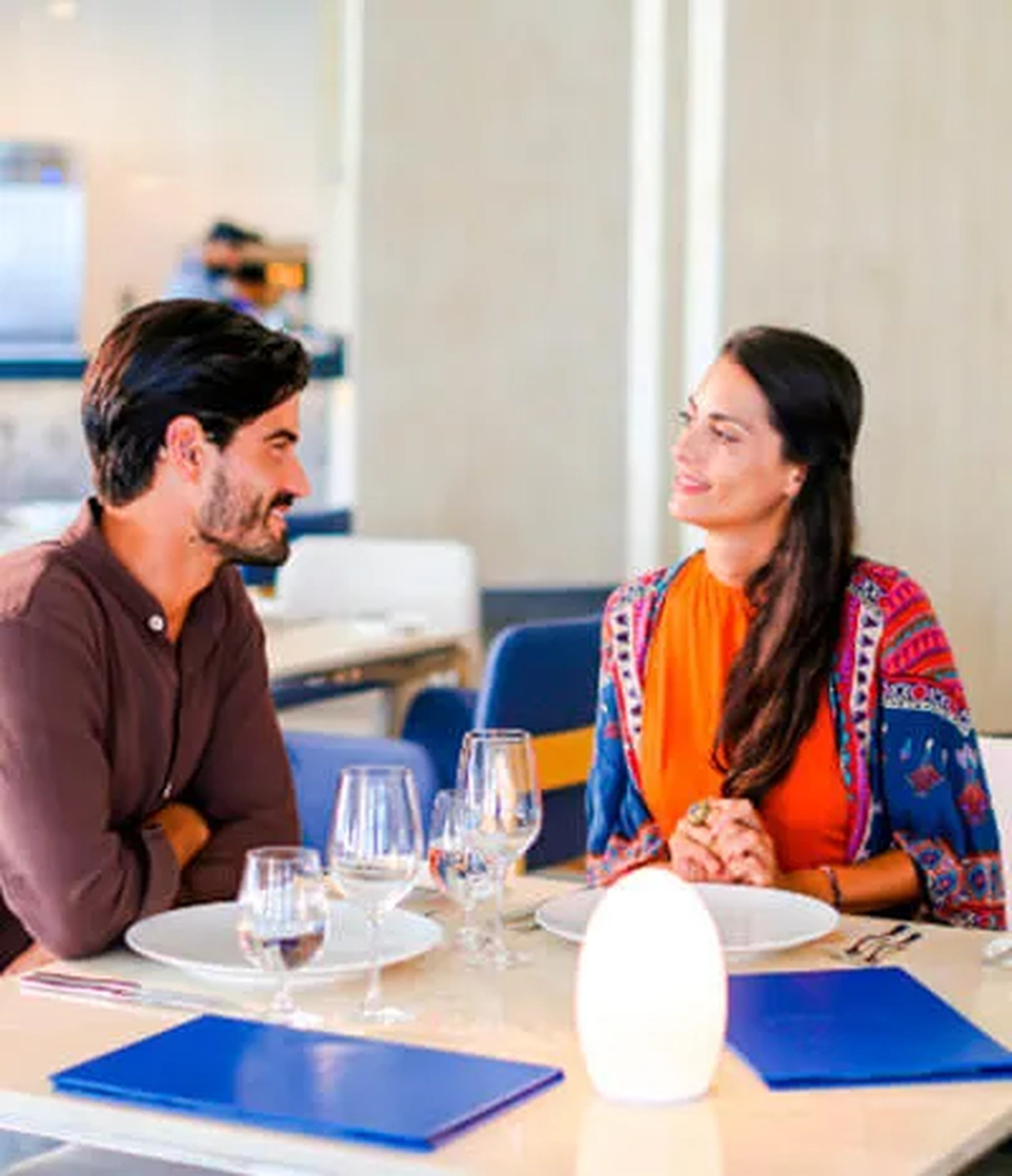 A man and woman sit at a restaurant table, smiling at each other, with glasses and plates set in front of them.
