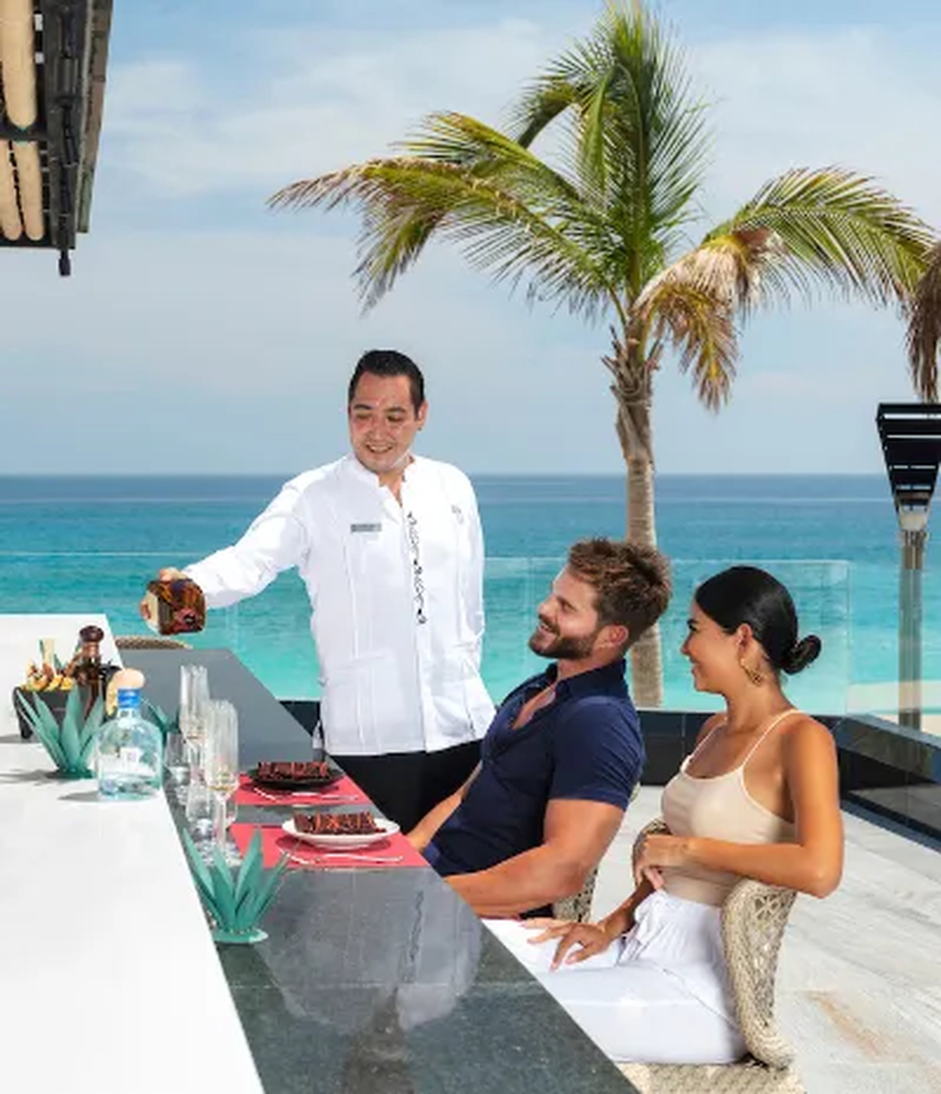 A waiter serves drinks to a smiling couple seated at an outdoor bar overlooking a tropical beach with palm trees and a clear blue sky.