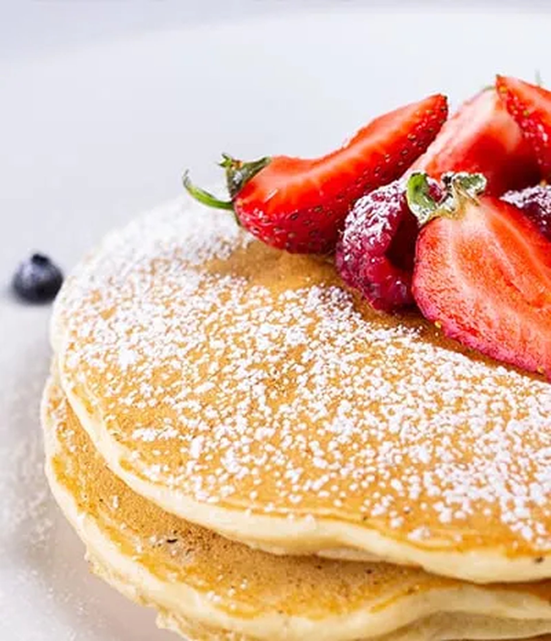 Stack of pancakes dusted with powdered sugar, topped with sliced strawberries and a blueberry on a white plate.