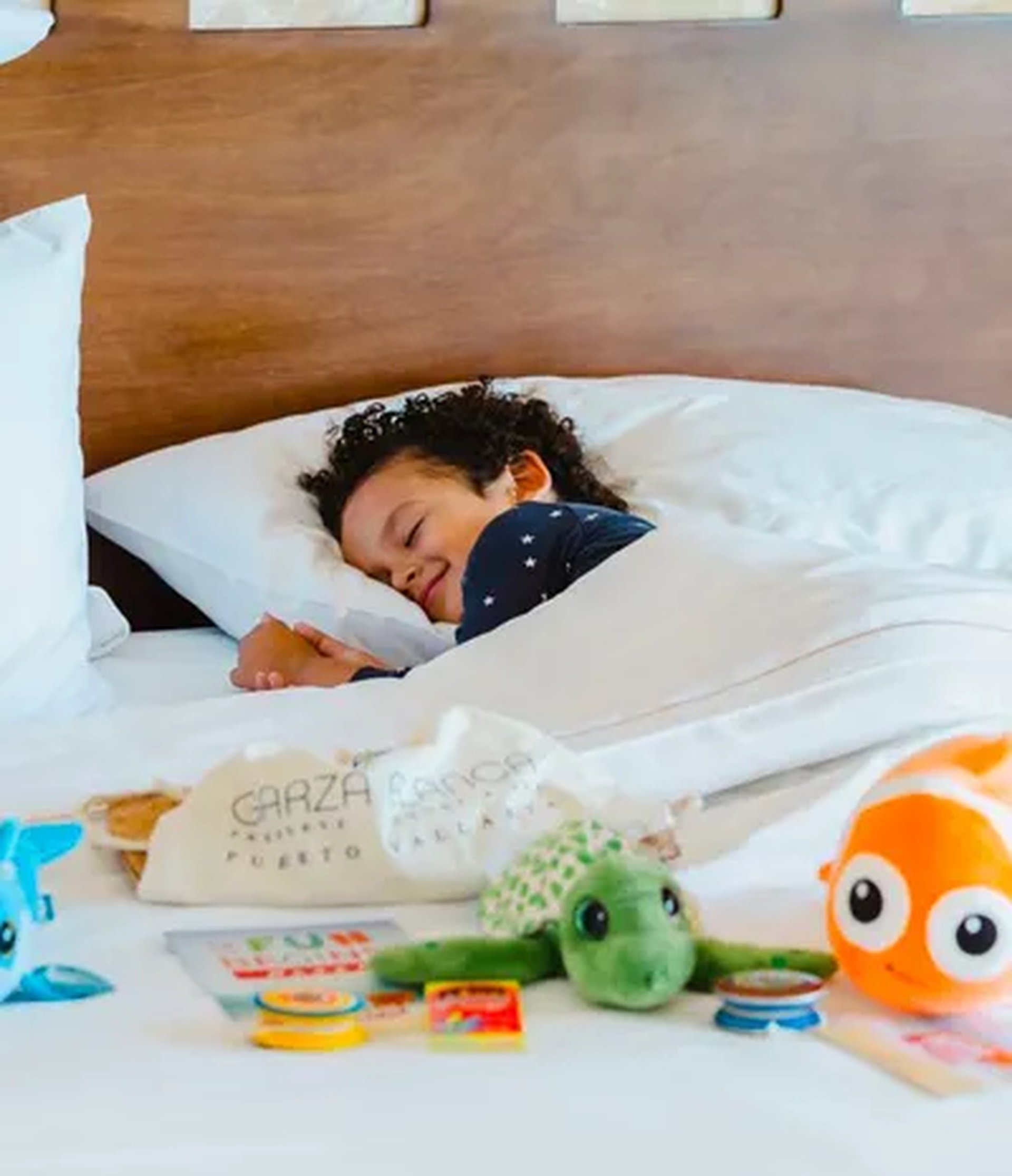 Child sleeping peacefully in bed with toys, including a turtle and orange fish, scattered on the blanket. Wooden headboard in the background.