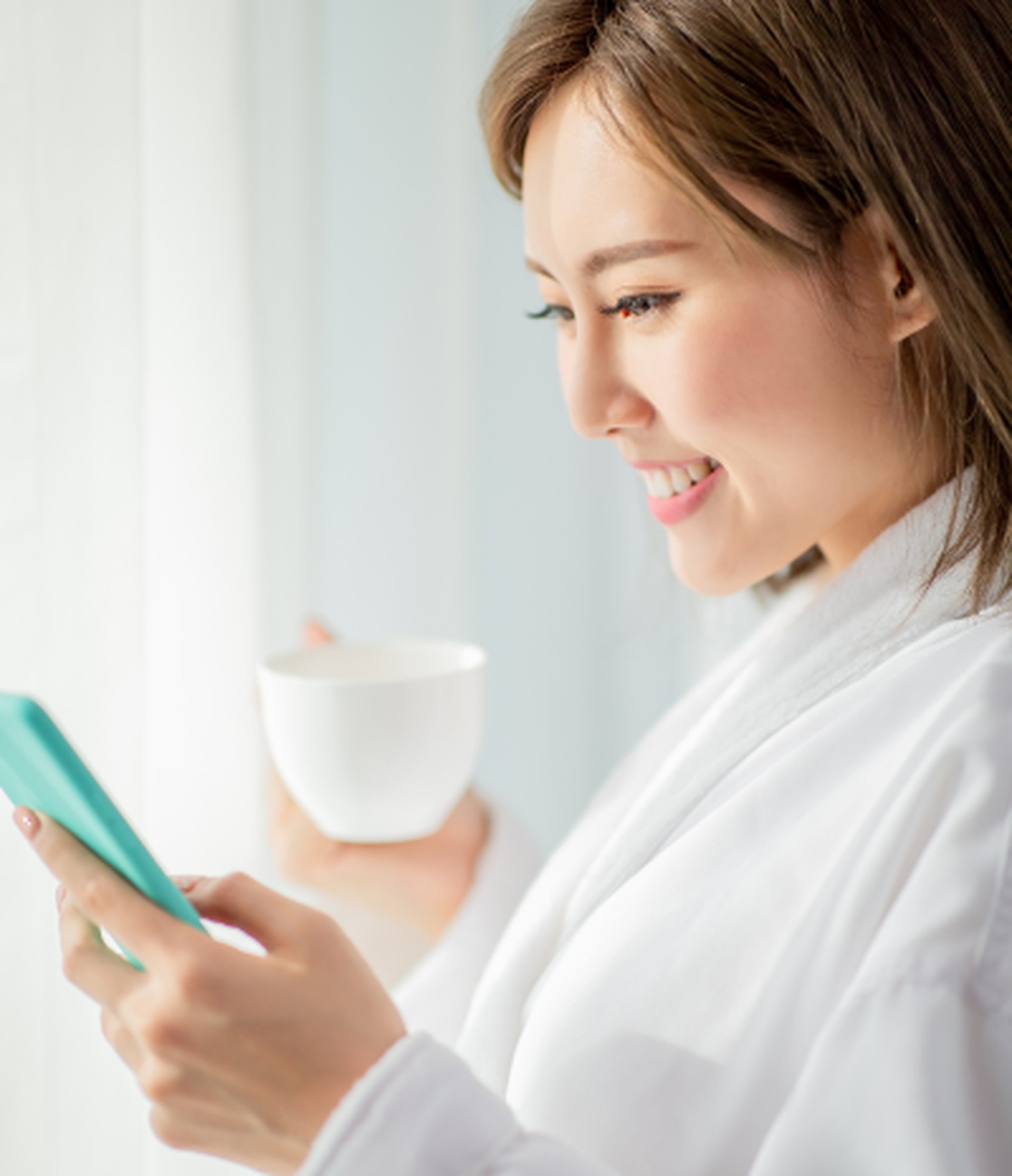 Woman in a white robe smiling at her phone while holding a white mug, standing near a window with soft light.