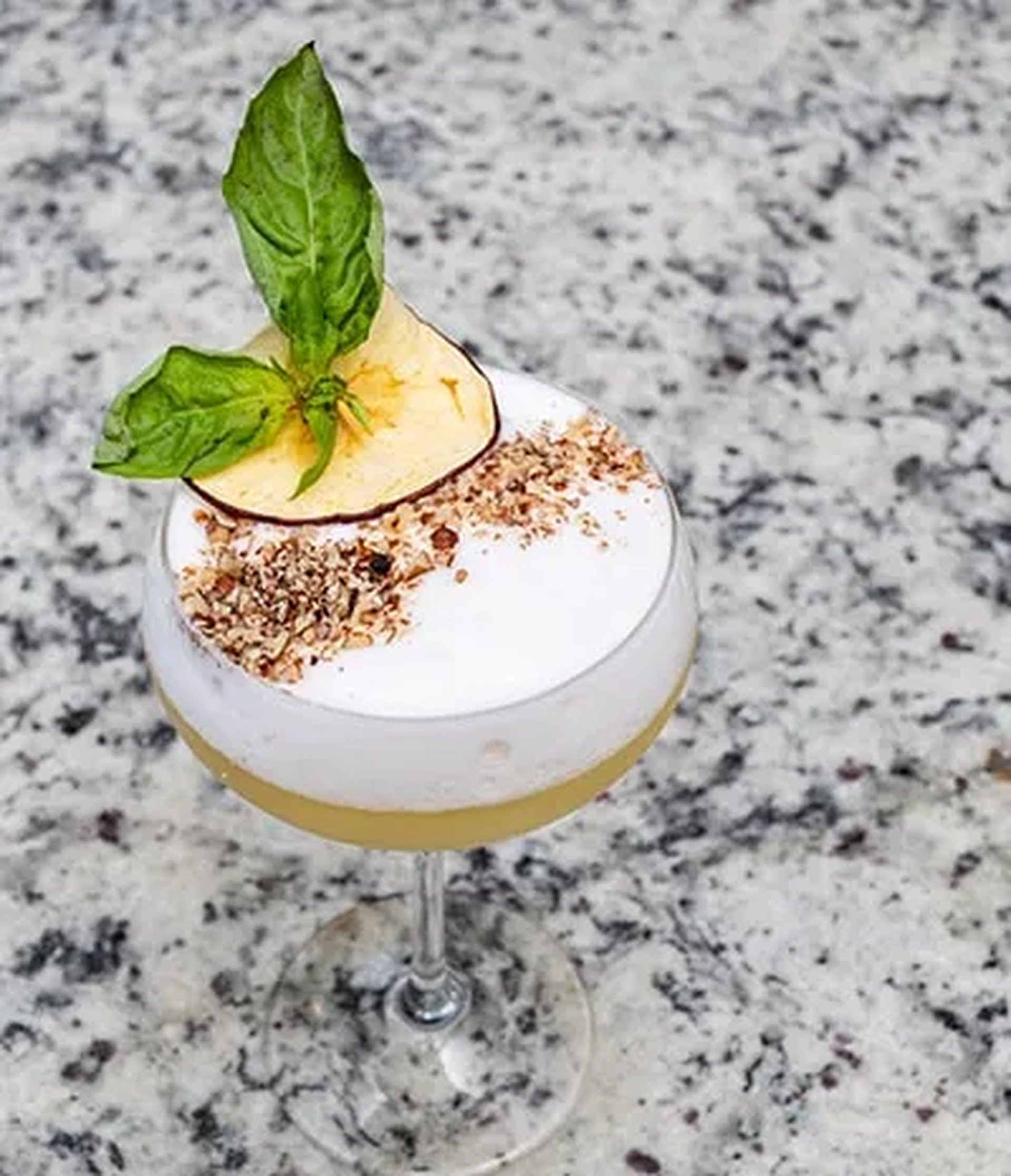 A cocktail with frothy white foam, topped with crushed nuts, a dried apple slice, and fresh basil, served in a stemmed glass on a granite surface.