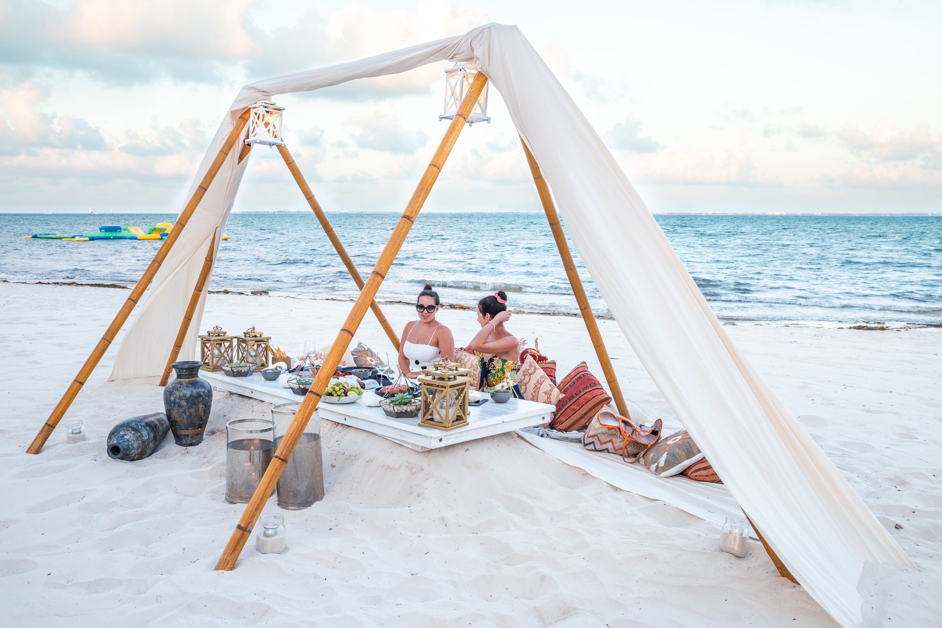 Two people sit under a canopy on a sandy beach, surrounded by cushions and decorative items, with the ocean in the background.