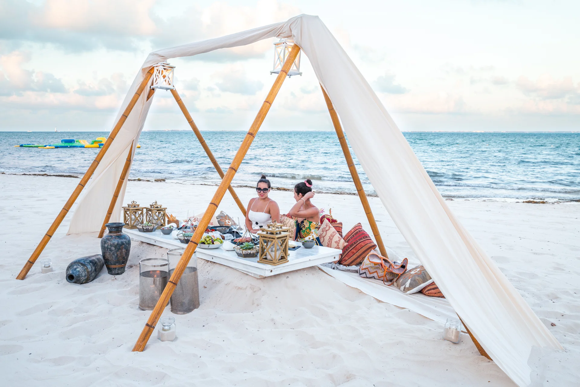 Two people sit under a canopy on a sandy beach, surrounded by cushions and decorative items, with the ocean in the background.