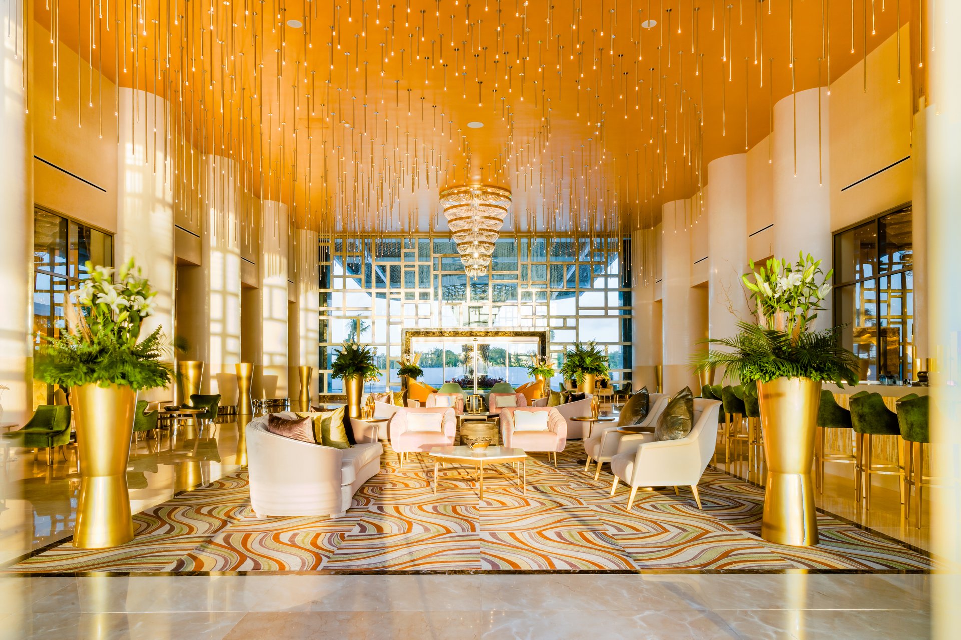 Luxurious hotel lobby with a golden ceiling, elegant chandelier, plush seating, vibrant plants, and a patterned carpet.