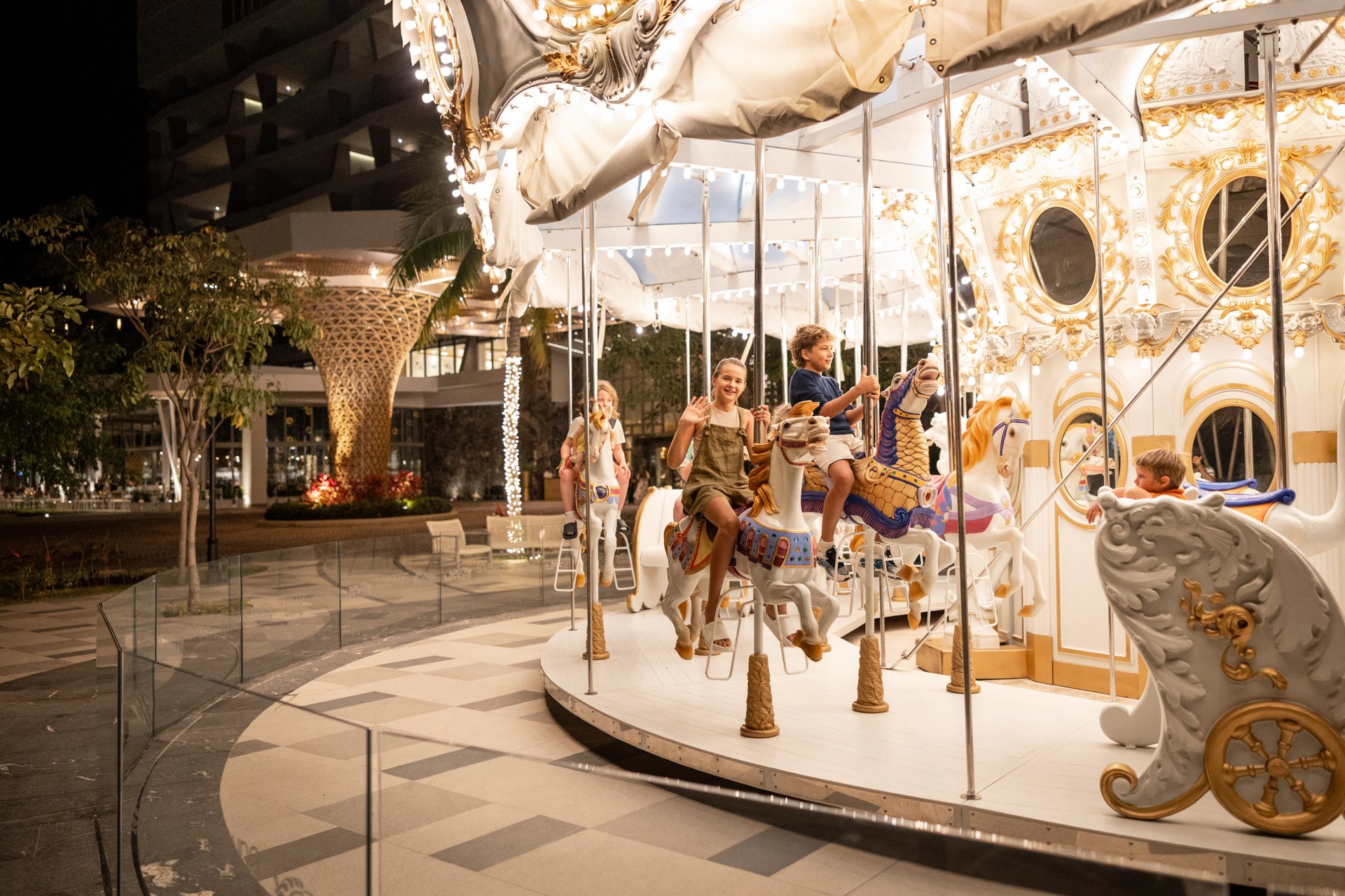 A family enjoys a nighttime carousel ride, with children on ornate horses and a lit-up background, in an outdoor setting.