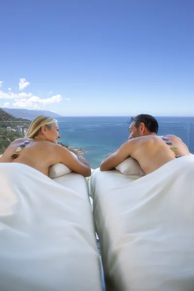 A couple enjoying a spa treatment with stones on their backs, overlooking a scenic ocean view under a clear blue sky.