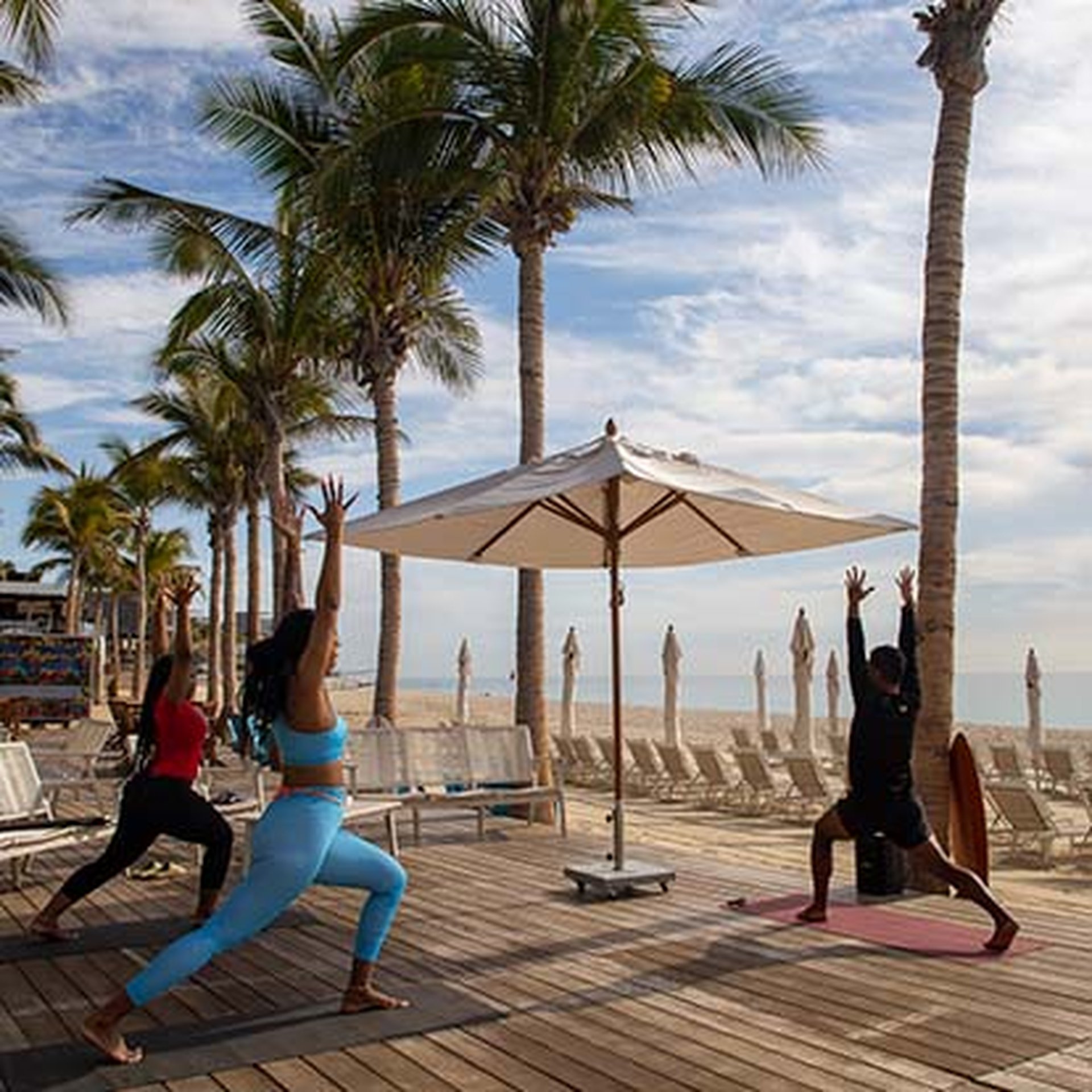 Three people practice yoga on a wooden deck by the beach, surrounded by palm trees and under a partly cloudy sky.