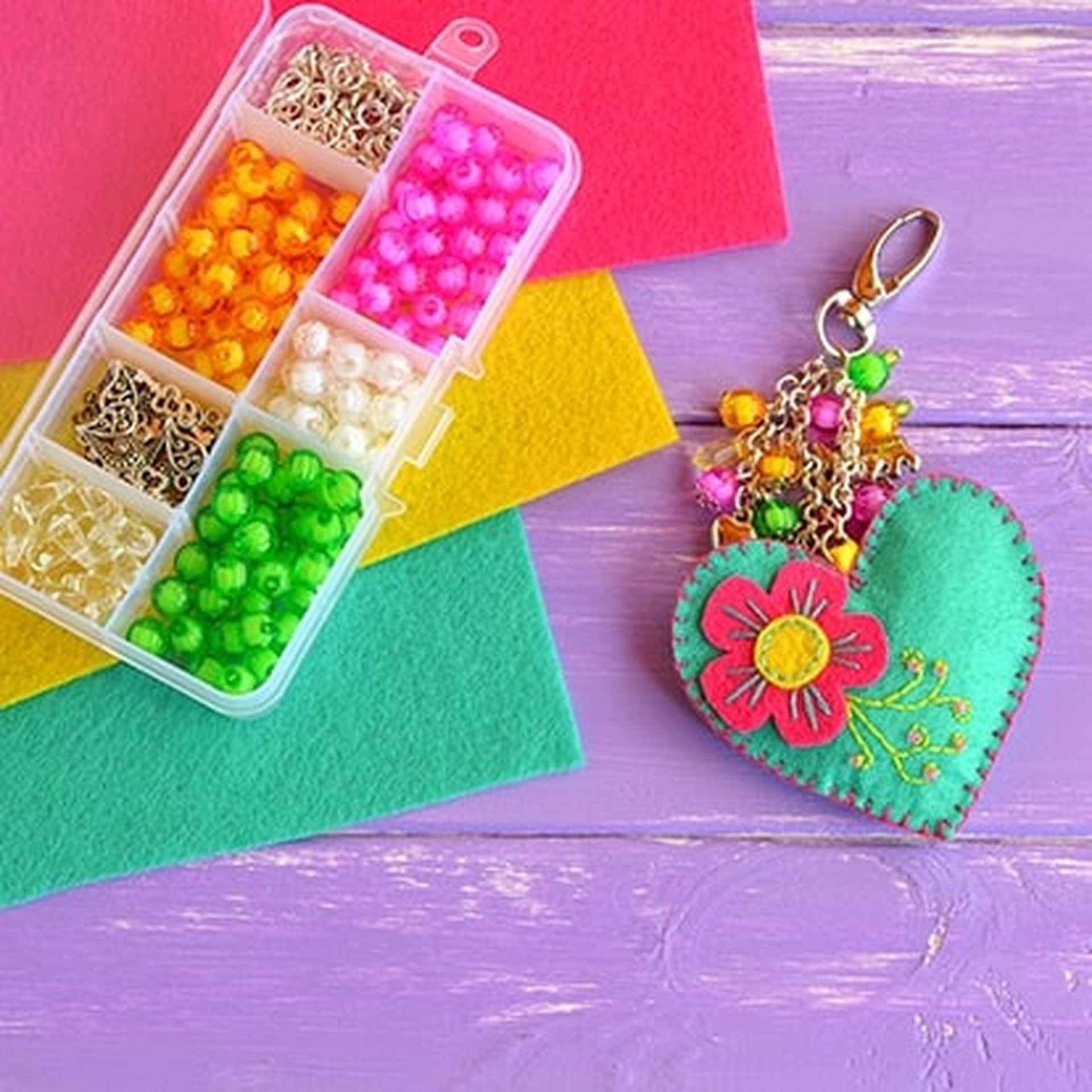 Colorful beads in a box and a handmade felt heart keychain with floral embroidery on a purple wooden surface with felt sheets.