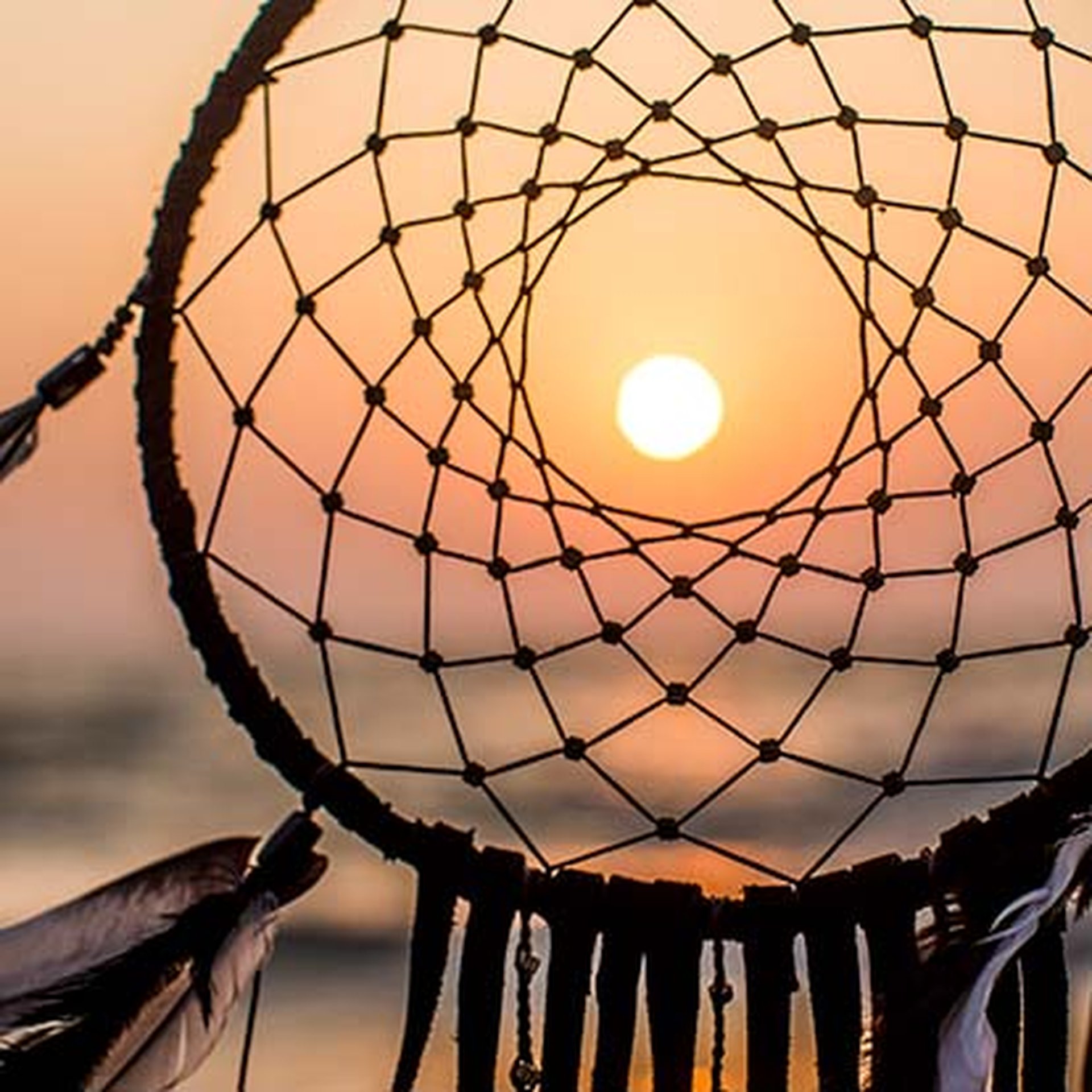 Dreamcatcher silhouetted against a vibrant sunset, with the sun perfectly centered in its web, over a blurred ocean background.