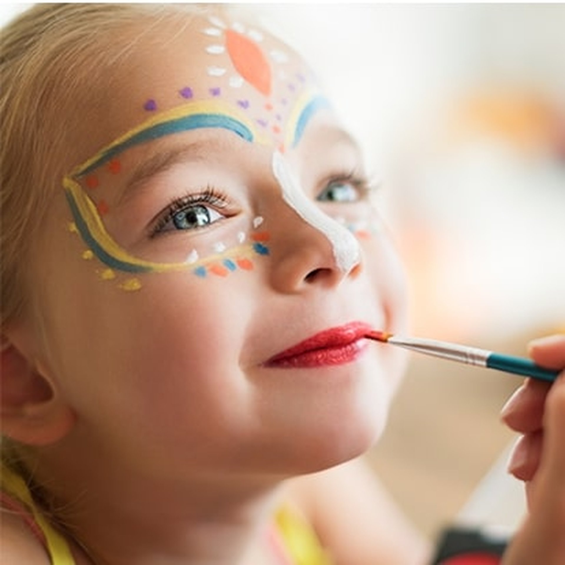 Child with colorful face paint, including dots and shapes, smiles while having lips painted red with a brush.