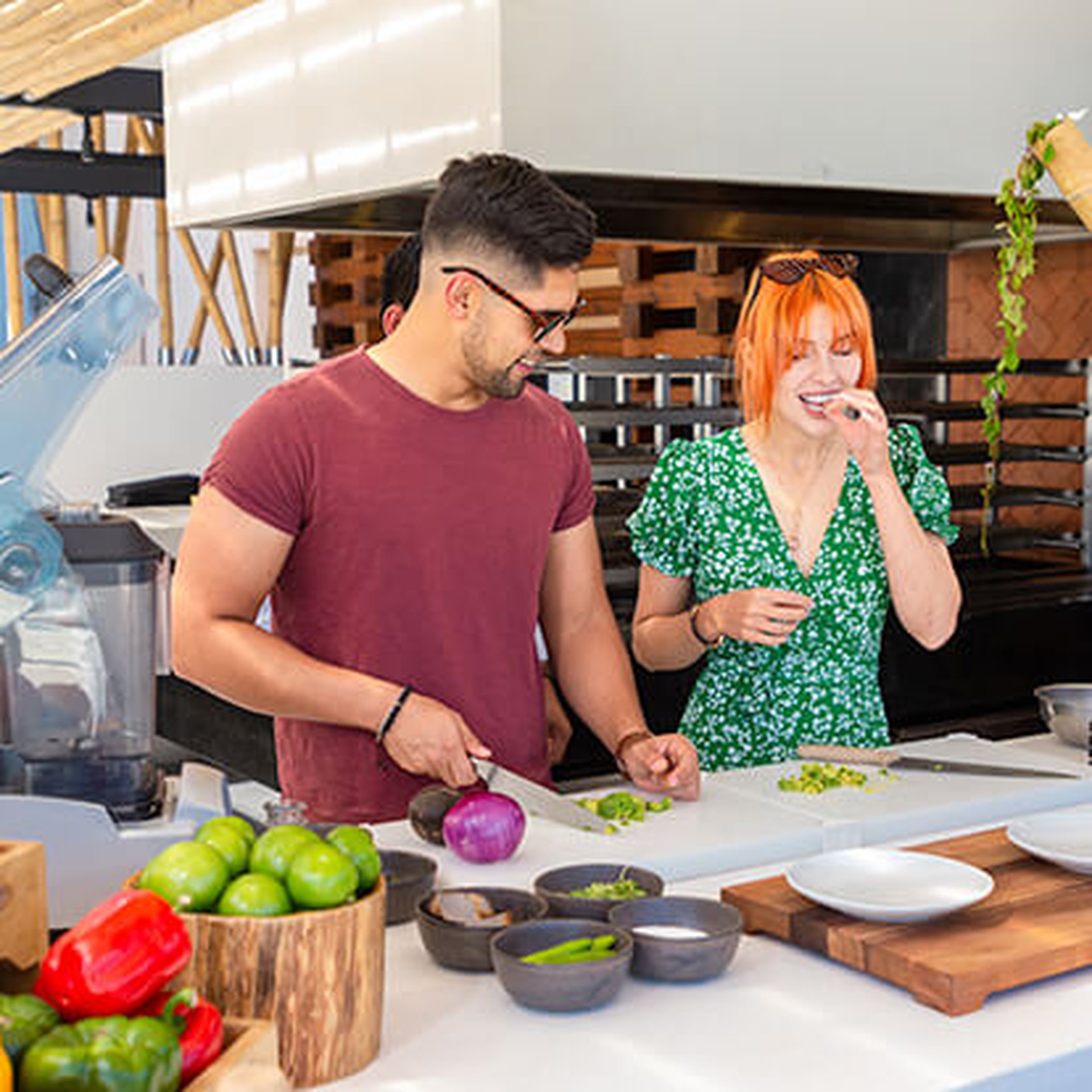 Two people in a kitchen, one chopping vegetables and the other smiling. Various fresh ingredients are on the counter.
