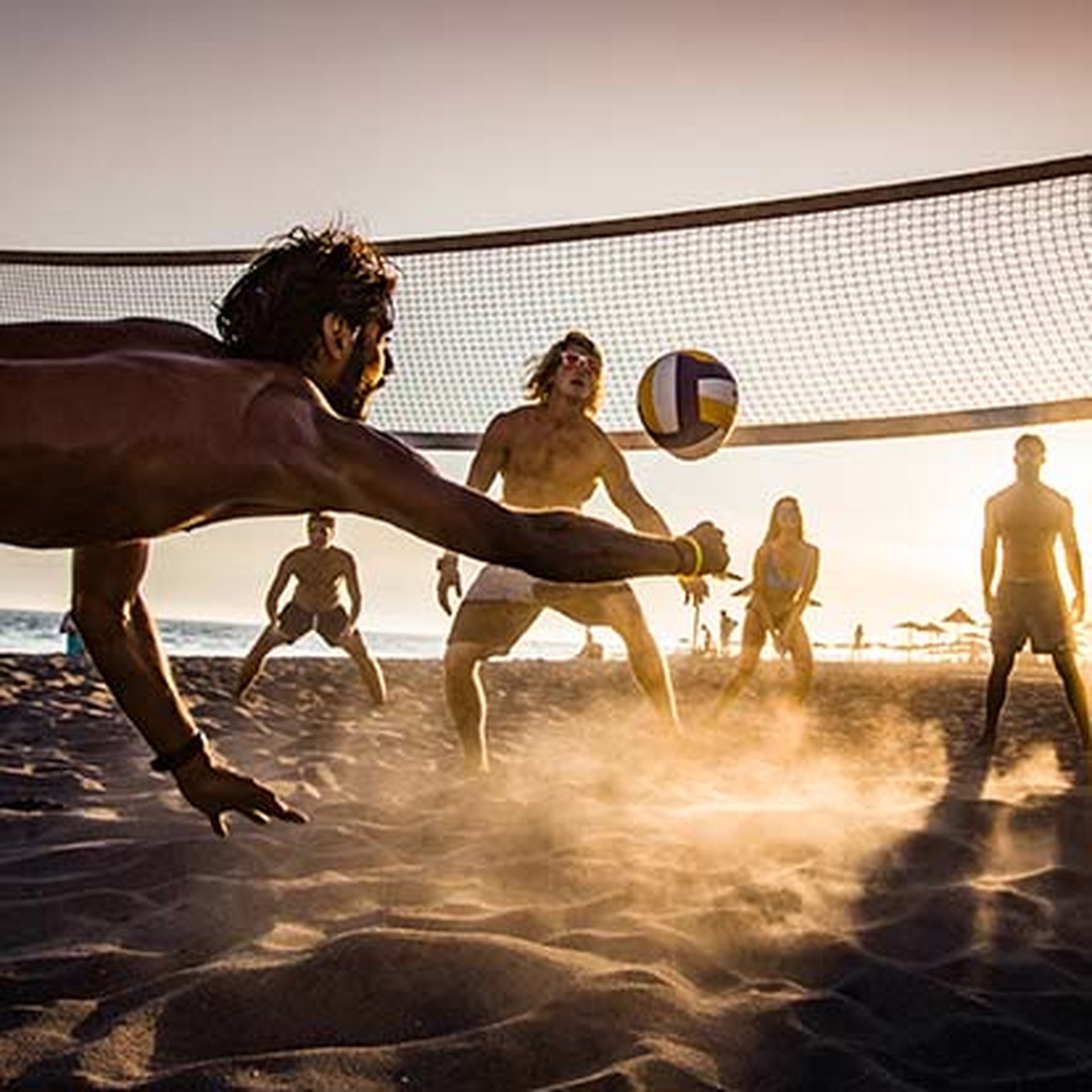 People playing beach volleyball at sunset, with a player diving for the ball as others watch. Sand is kicked up, and a net is visible.