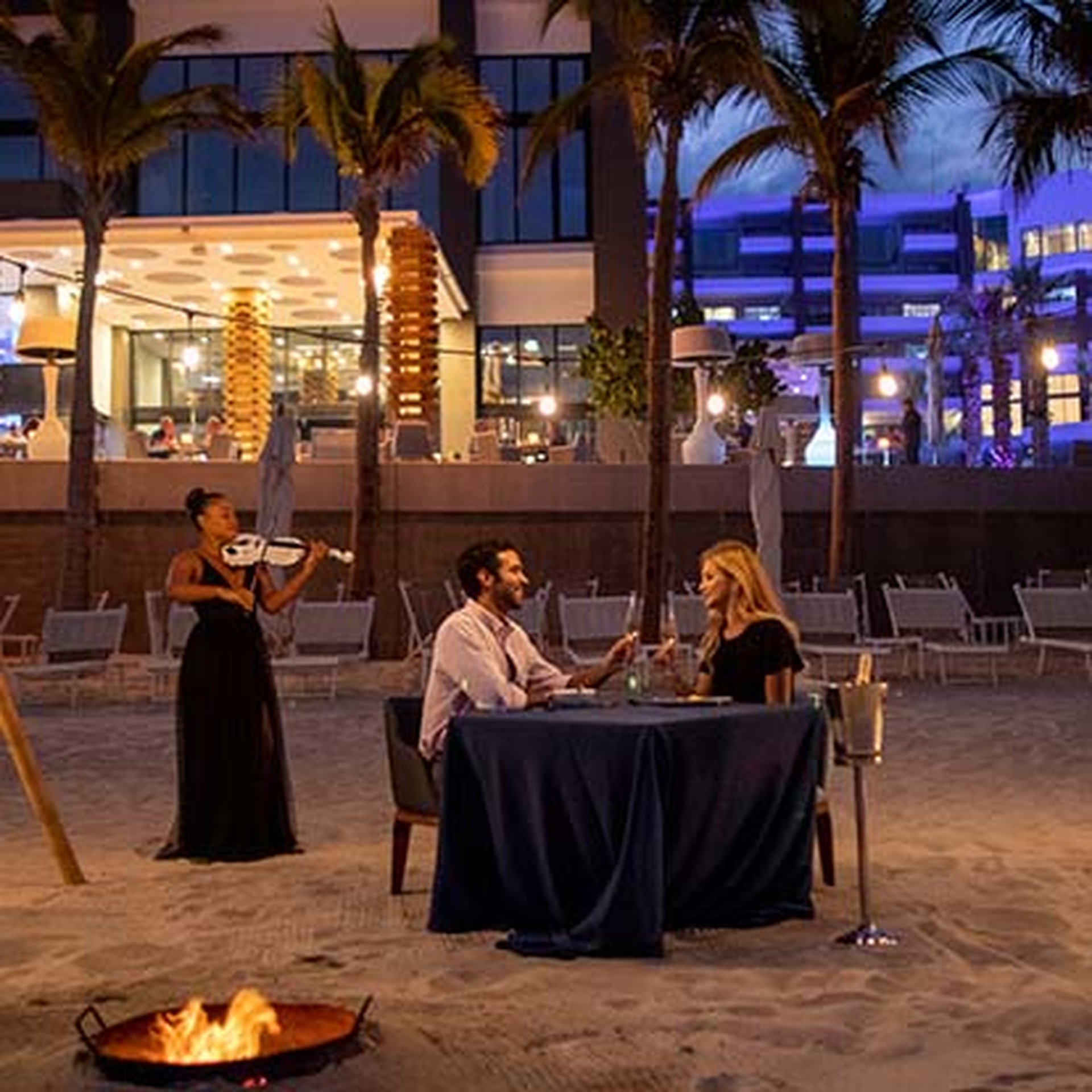 A couple enjoys a candlelit dinner on a beach at night, with a violinist playing nearby and a fire pit glowing in the foreground.