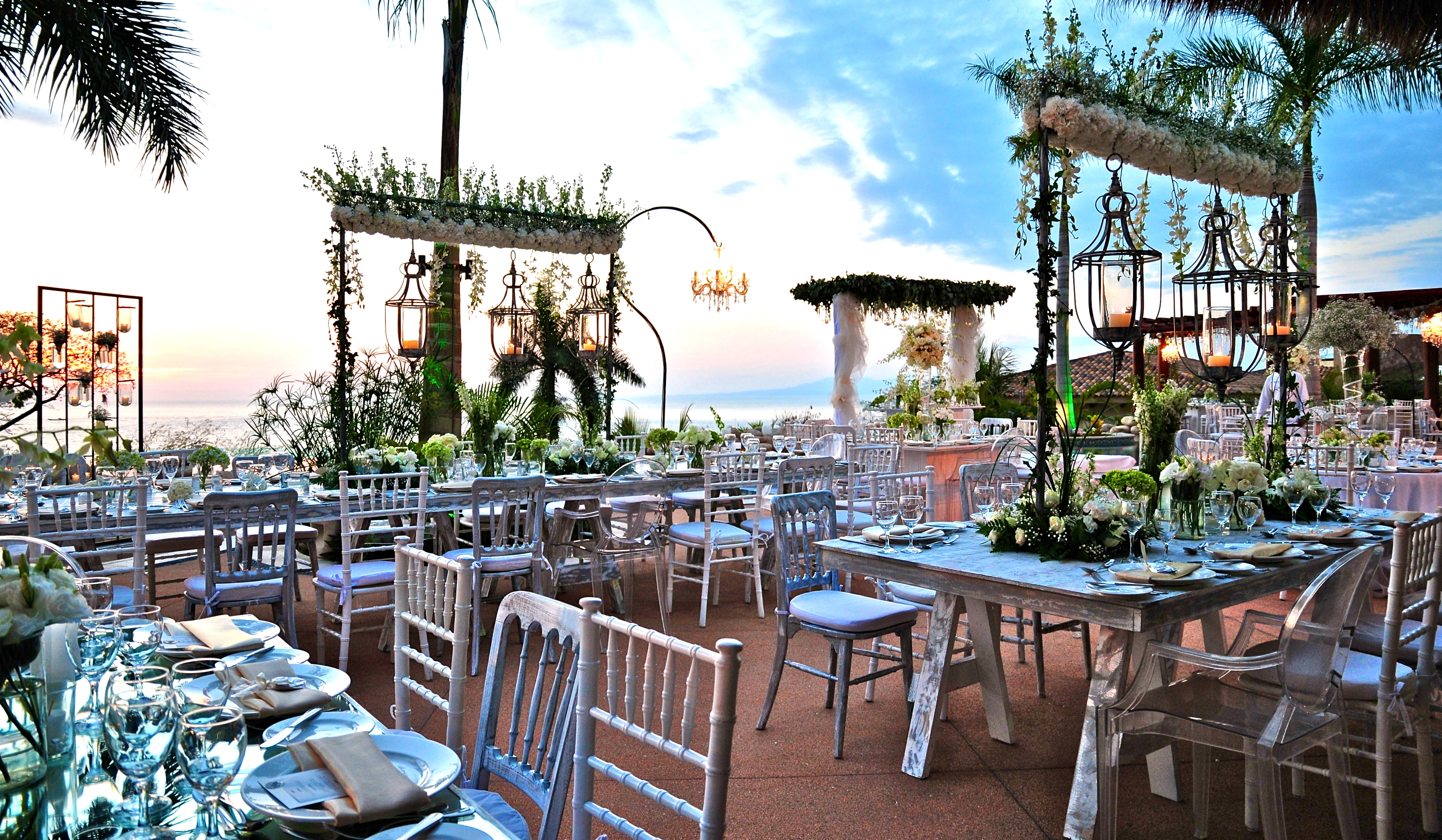 Elegant beachfront wedding setup with white floral decor, lanterns, and a stunning ocean view at sunset.