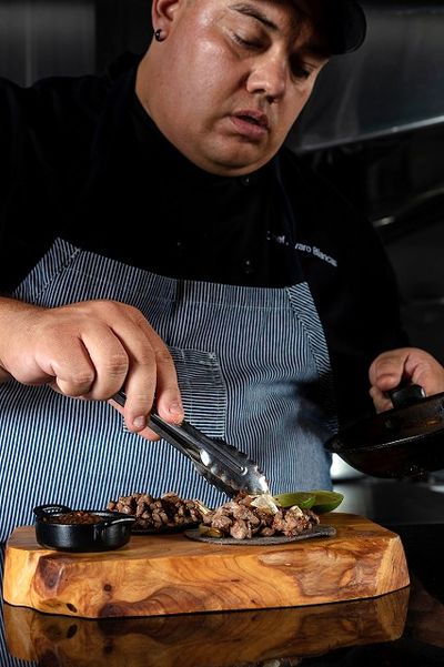 Chef plating cooked meat on a wooden board with tongs, wearing a striped apron and black shirt, in a kitchen setting.