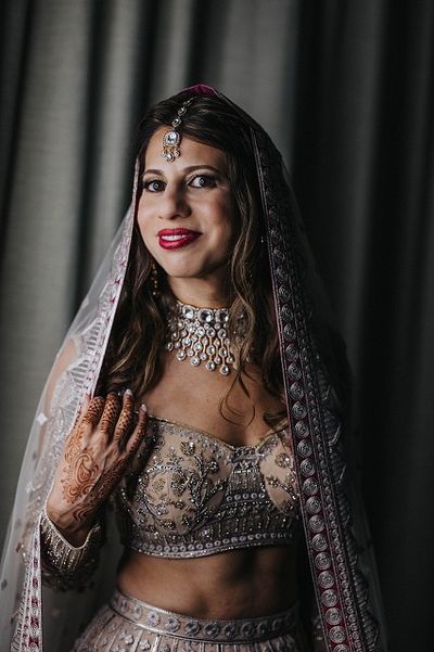 A woman in an ornate bridal outfit with intricate jewelry and henna, smiling softly, stands against a dark curtain backdrop.