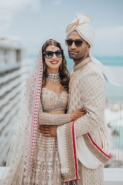 A couple in traditional Indian wedding attire, wearing sunglasses, stands together with a scenic ocean backdrop.