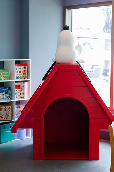 A Snoopy statue sits atop a large red doghouse inside a room with bookshelves filled with children's books.