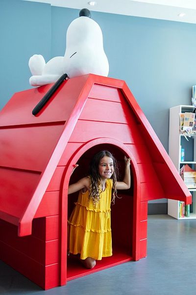 A girl in a yellow dress kneels inside a red doghouse with a Snoopy figure on top, against a blue wall background.