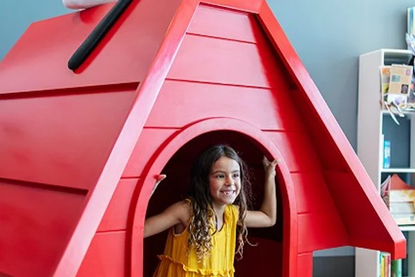 A girl in a yellow dress kneels inside a red doghouse with a Snoopy figure on top, against a blue wall background.