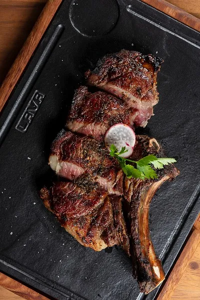 Grilled tomahawk steak on a black slate, garnished with a dollop of butter and parsley, served on a wooden board.