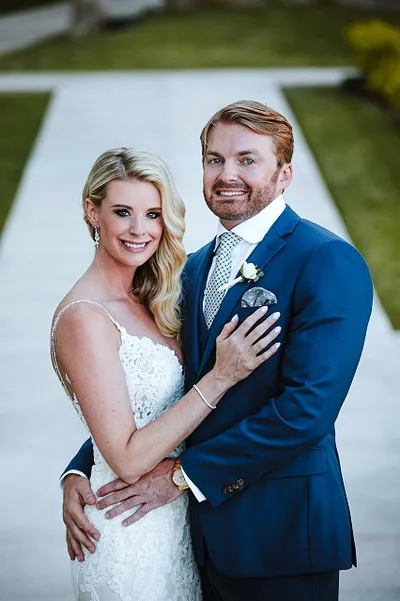 Bride in a white lace dress and groom in a blue suit smiling and embracing outdoors on a stone pathway.