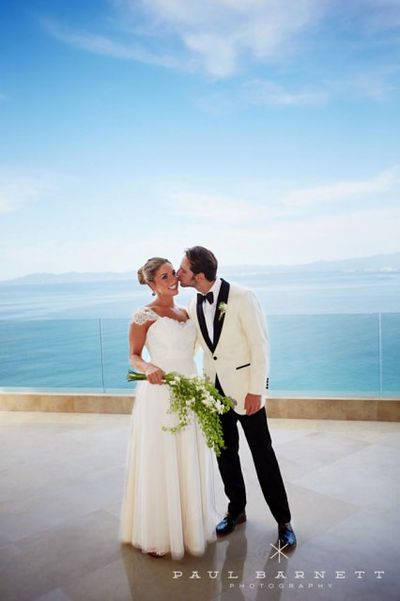 A couple in wedding attire shares a kiss on a balcony overlooking the ocean under a clear blue sky.
