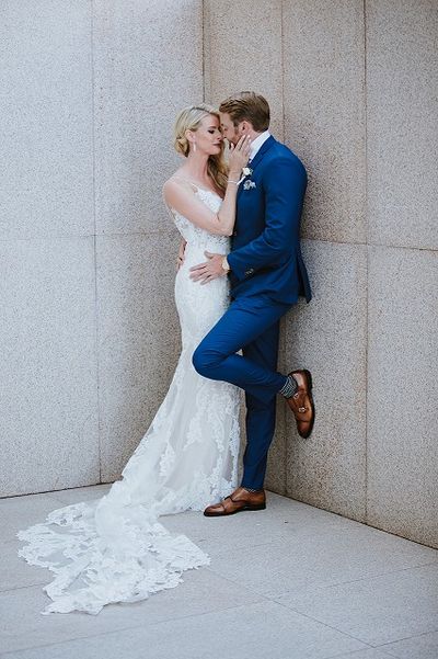 Bride in a lace gown and groom in a blue suit share an intimate moment against a stone wall.