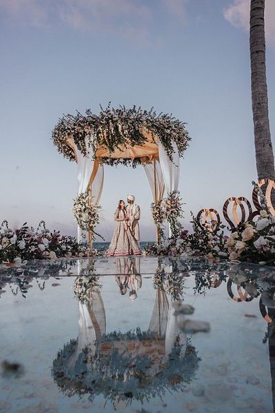 A couple in elegant attire stands under a floral arch by the ocean, reflected in a glassy surface, with a clear blue sky above.