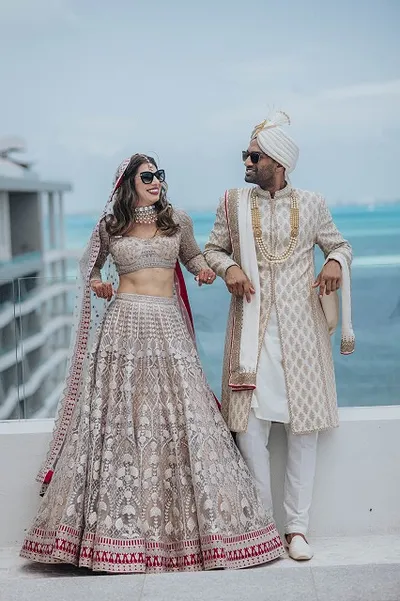 A joyful couple in traditional attire, wearing sunglasses, stands on a balcony with the ocean in the background.