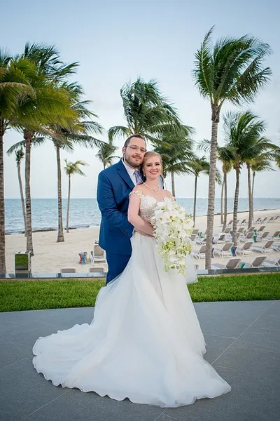 A newlywed couple poses on a beachfront terrace, surrounded by palm trees, with the ocean in the background.