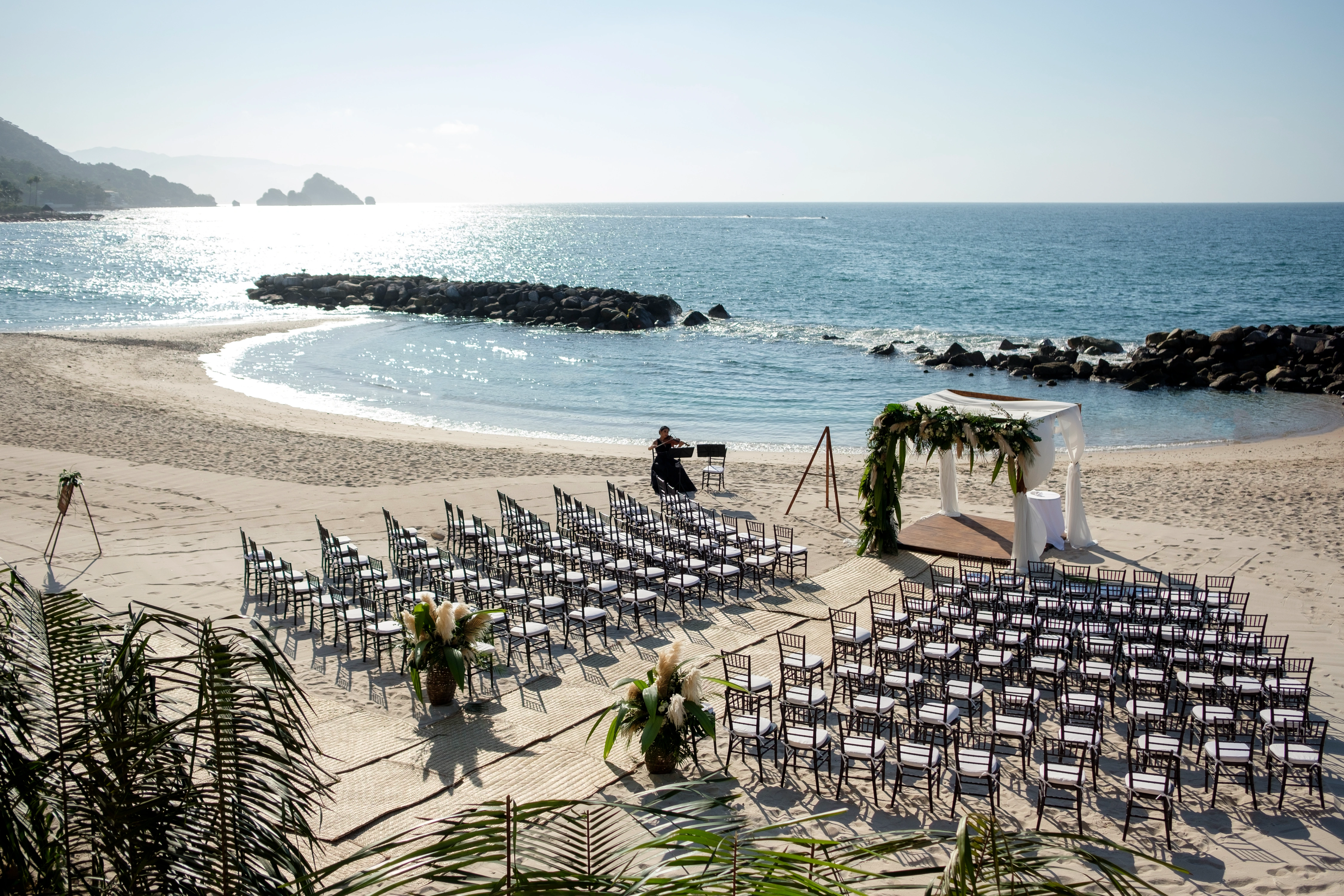 Wedding setup on a pristine beach with arranged chairs facing a floral arch by the sea under a clear blue sky.