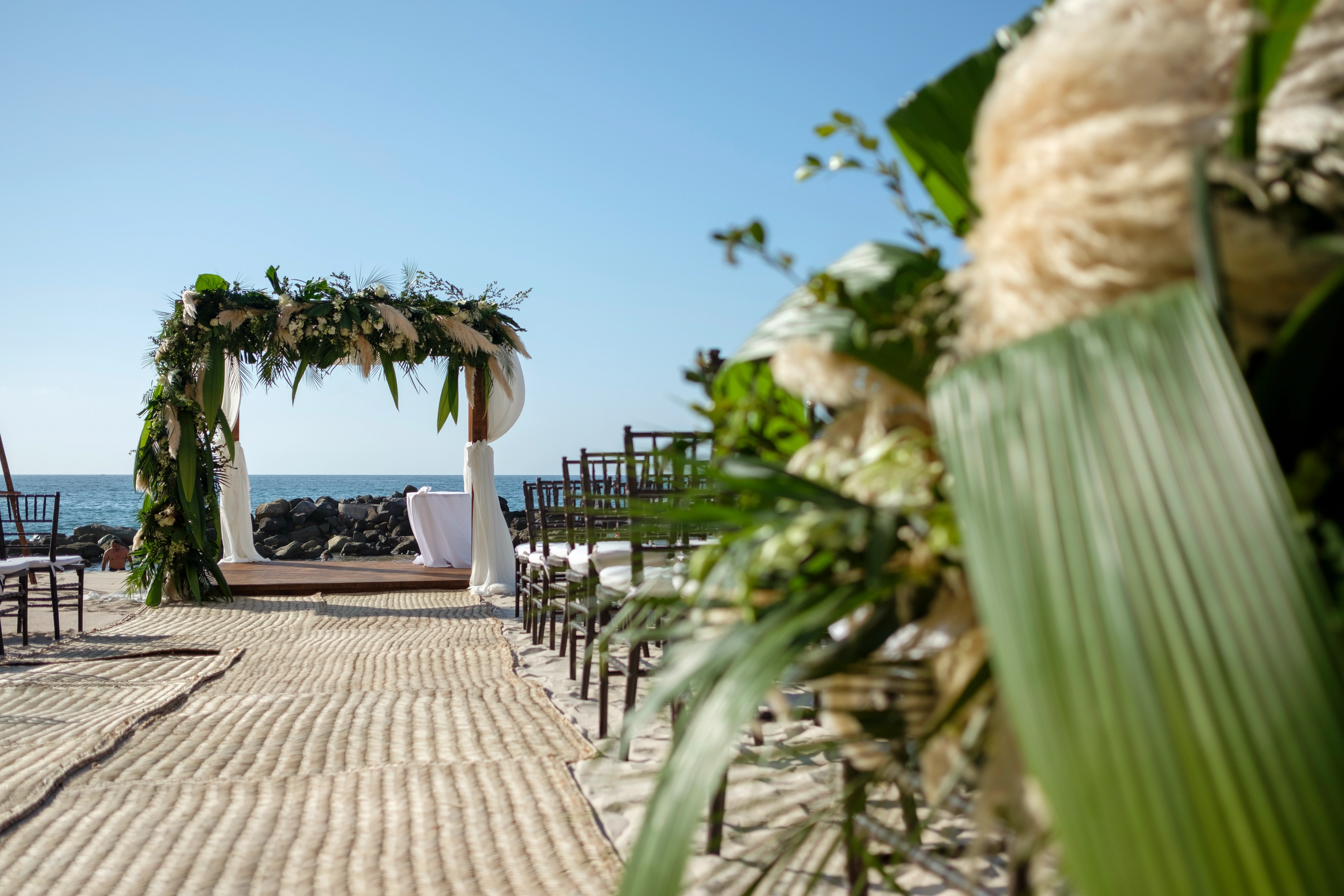 Beachfront wedding setup with a floral arch, wooden chairs, and a woven aisle, overlooking the ocean under a clear blue sky.