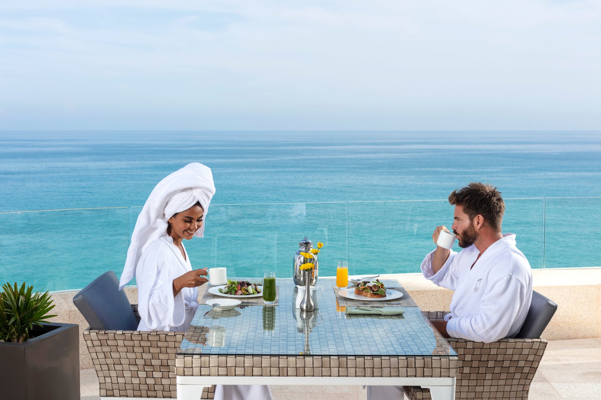 A man and woman in bathrobes enjoy breakfast at a seaside table, with ocean views, fresh juice, and coffee.