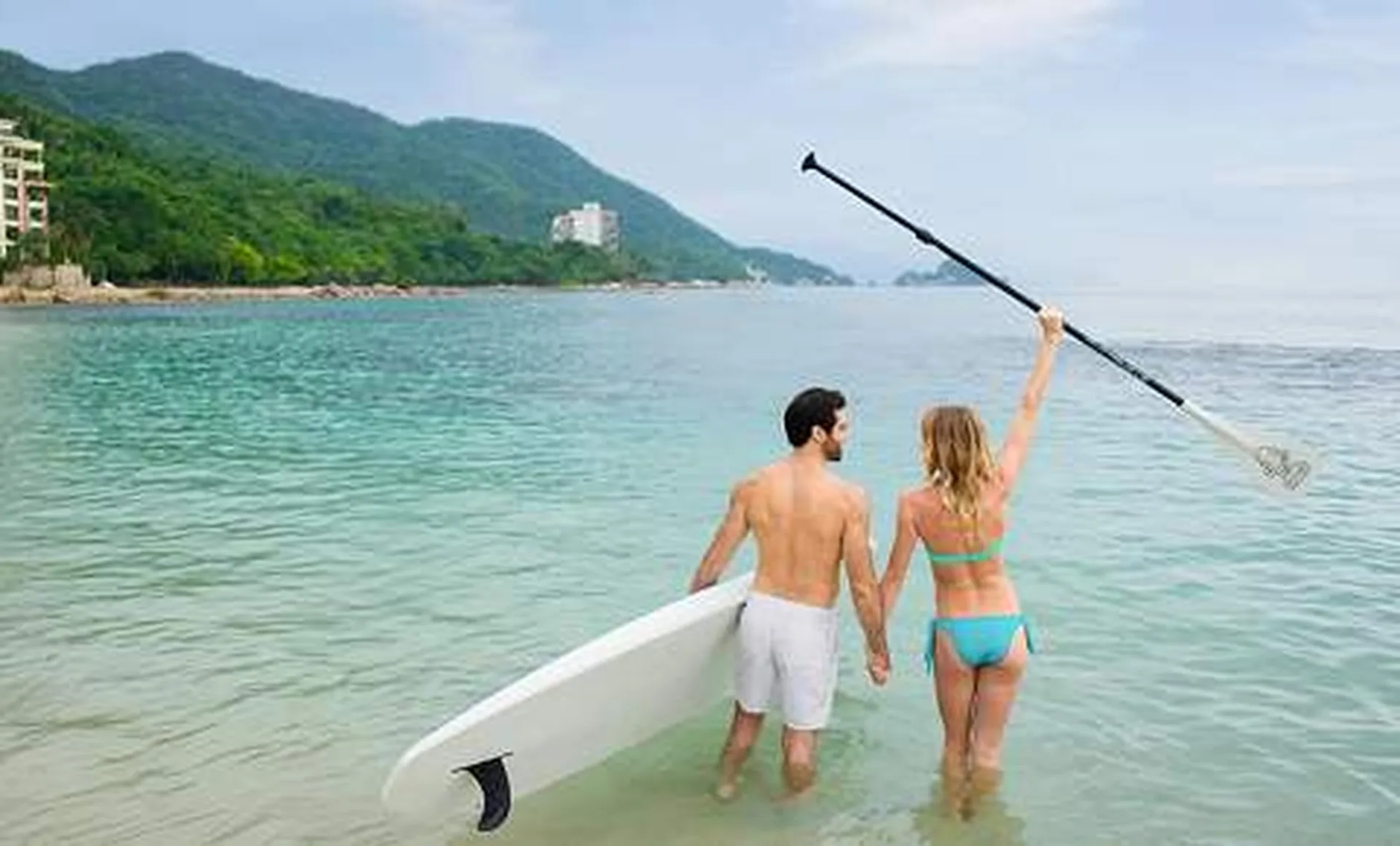 A couple stands on a beach, holding a paddleboard and paddle, facing the ocean with lush green hills in the background.