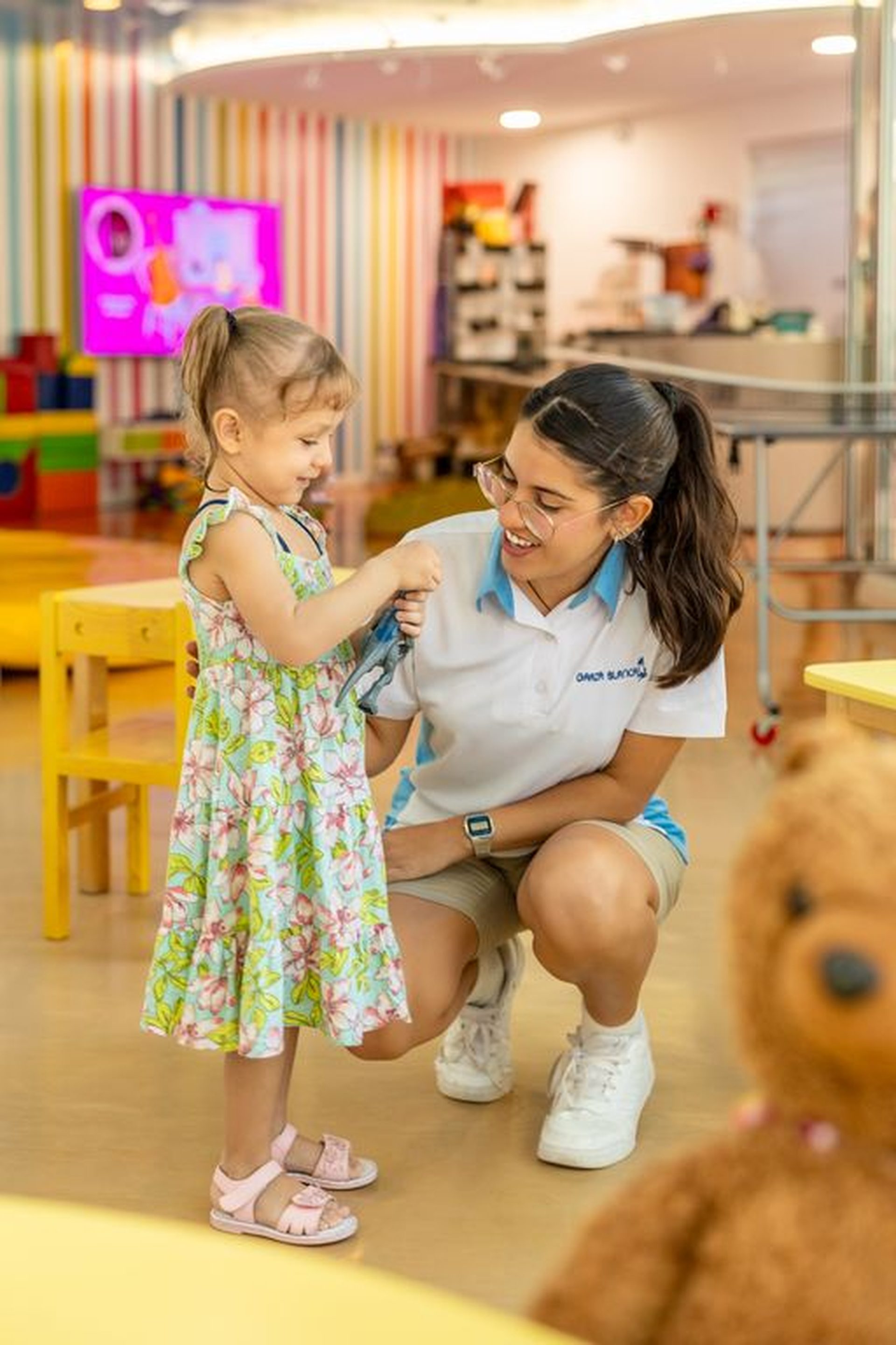 A smiling resort staff member kneels to engage with a young girl in a colorful playroom.